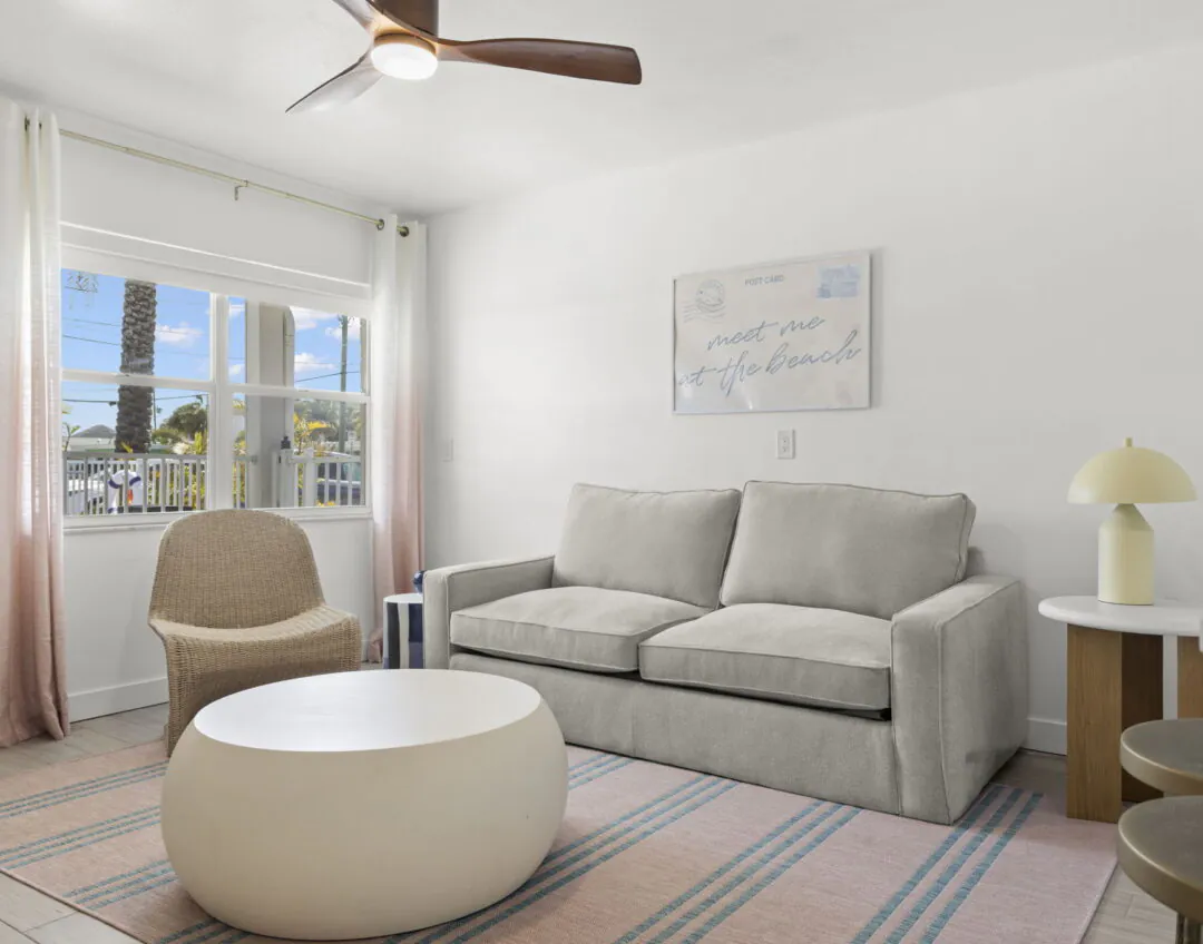 Bright living room at Hotel Cabana with gray sofa, wicker chair, round coffee table, white curtains, and a wall sign that says meet me at the beach.