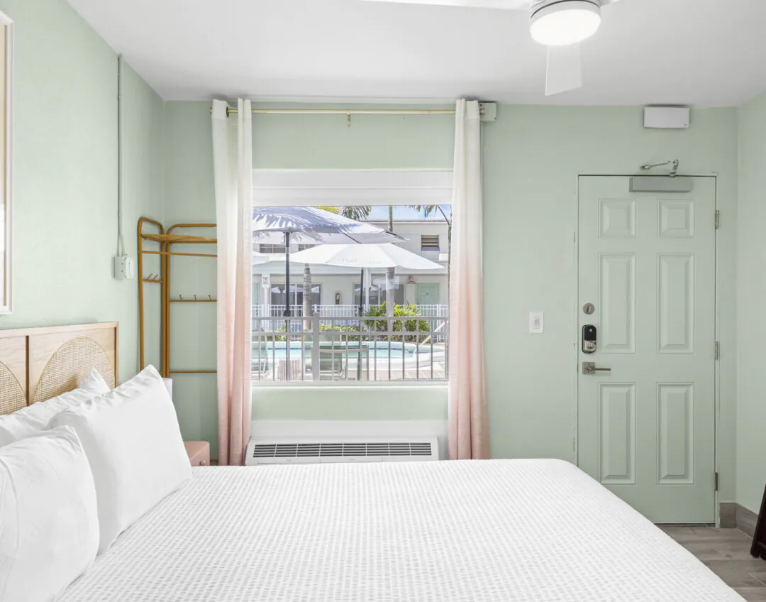 A bright Hotel Cabana room with a bed, pastel green walls, and a window overlooking the pool area with umbrellas and lounge chairs outside.
