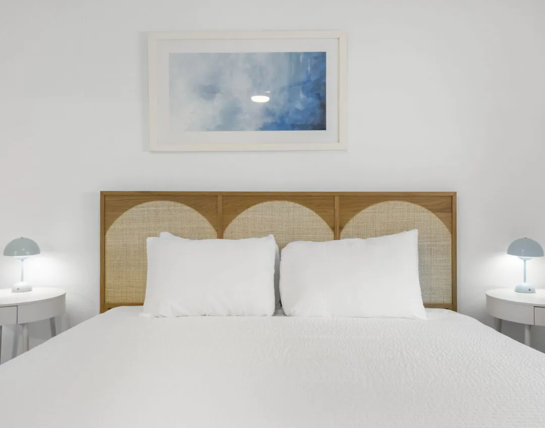 Minimalist Hotel Cabana bedroom with a bed, rattan headboard, two white pillows, side tables, blue lamps, and an abstract painting on a white wall.