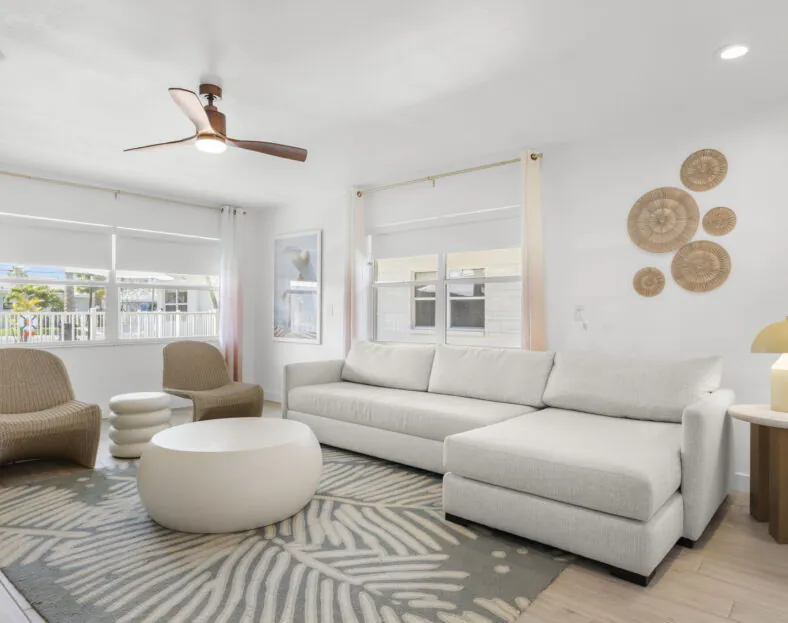 Chic Hotel Cabana living room with a white sectional sofa, round coffee table, wicker chairs, wall art, and large windows with white curtains.