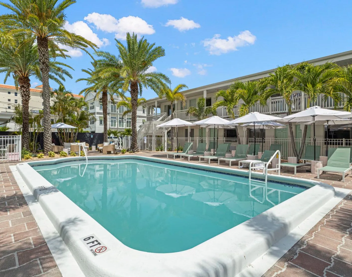 Hotel Cabana’s 90-foot rectangular outdoor pool is surrounded by lounge chairs, umbrellas, palm trees, and a two-story building under a blue sky with clouds.
