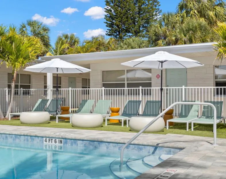 Hotel Cabana’s outdoor pool, lined with white umbrellas, lounge chairs, and palm trees, sits before a one-story building beneath a blue sky.